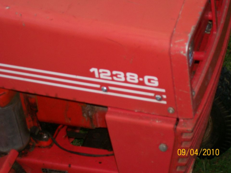 Red garden tractor hood labeled 1238-G with white stripes, weathered paint, on grass