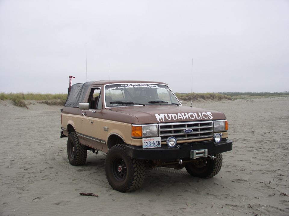 Lifted brown and tan Ford Bronco with 'Mudaholics' hood lettering parked on a sandy beach