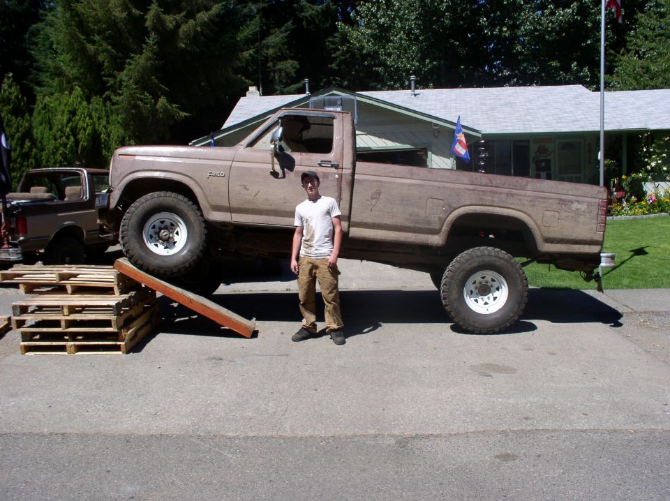 Brown Ford F-250 pickup with front wheel up on a pallet ramp, man standing beside it; Bronco in background