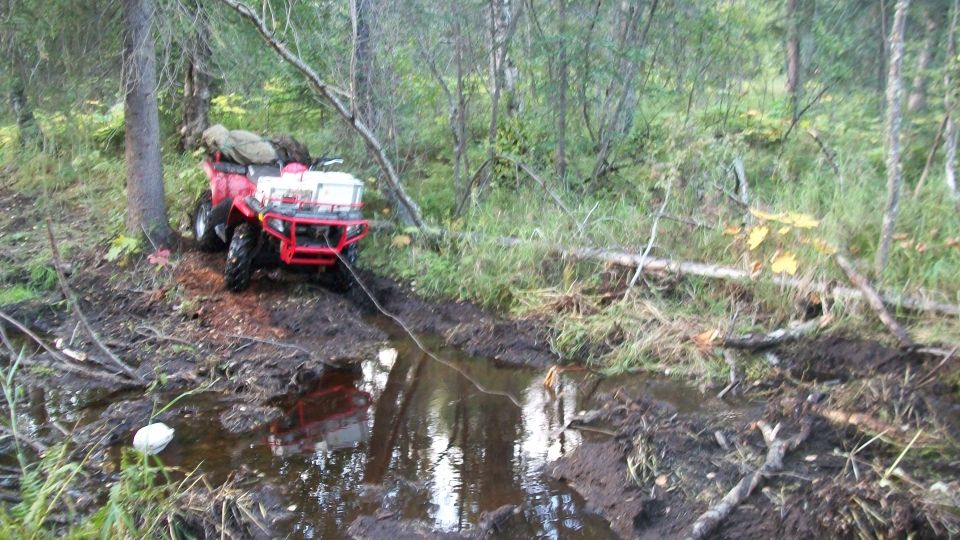 Red ATV stuck in a muddy ditch in the woods, with a rope attached