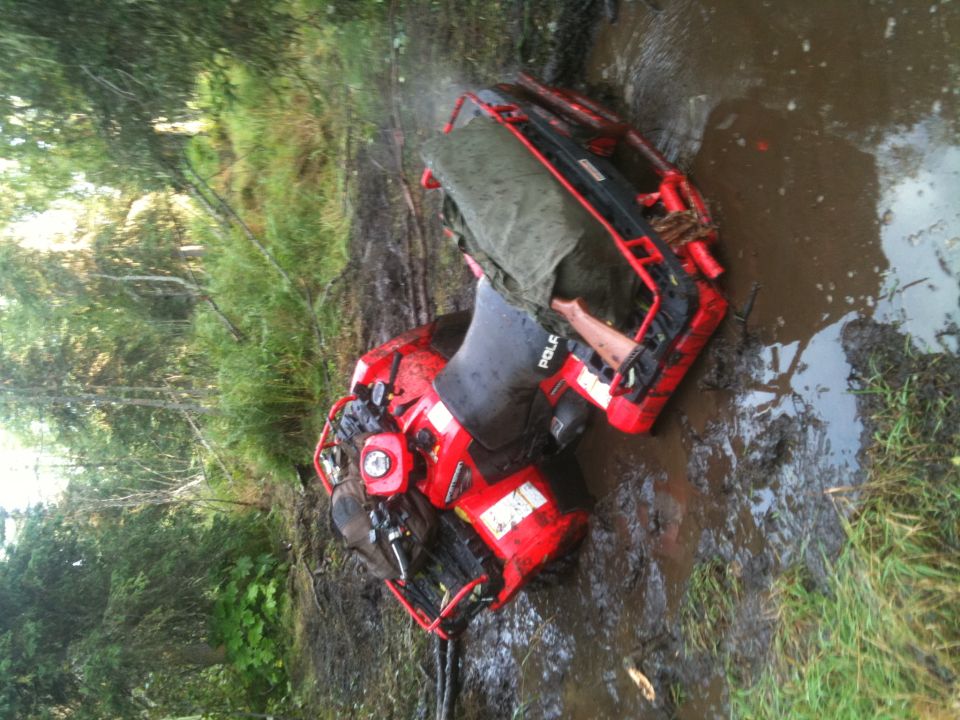 Red ATV stuck in a muddy ditch with trees in the background. Text on the side reads 'POLARIS'