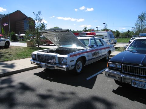 1991 Ford LTD Crown Victoria - white police sedan with red stripes and an open hood sits next to a black sedan marked POLICE at an outdoor car show.
