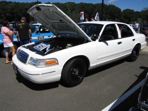 1991 Ford LTD Crown Victoria - white Ford Crown Victoria police-style sedan with its hood raised showing the engine, displayed among other vehicles at a car show.