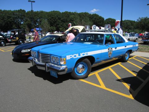 1991 Ford LTD Crown Victoria - light blue vintage four-door police sedan lettered 'Police' with a shield decal and roof lights, parked beside a dark car at a show.