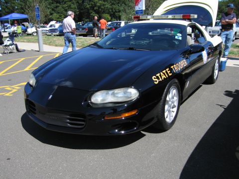 1991 Ford LTD Crown Victoria - black Chevrolet Camaro lettered 'State Trooper' in gold with a roof light bar, parked on asphalt at a car show.