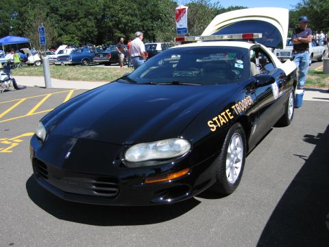 1991 Ford LTD Crown Victoria - black Chevrolet Camaro with gold 'State Trooper' lettering and roof beacons displayed among other vehicles at an outdoor show.