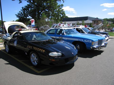 1991 Ford LTD Crown Victoria - black 'State Trooper' Chevrolet Camaro parked next to a light blue 'Police' sedan, both with roof lights, at a sunny car show.