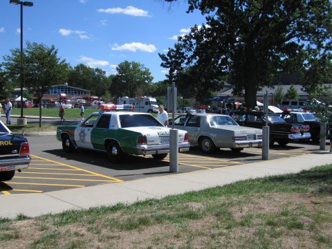 1991 Ford LTD Crown Victoria - green and white police sedan, a tan sedan and other patrol cars parked in a striped lot with show crowds in the background.