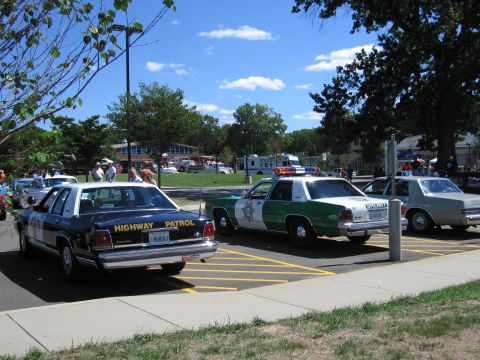 1991 Ford LTD Crown Victoria - Several vintage police sedans, including a dark 'Highway Patrol' car and a green-and-white sheriff car, lined up in a striped lot.