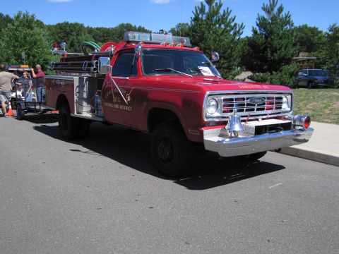 1991 Ford LTD Crown Victoria - red lifted 4x4 fire engine with chrome grille and roof lights, towing a small trailer, parked on pavement among show vehicles.