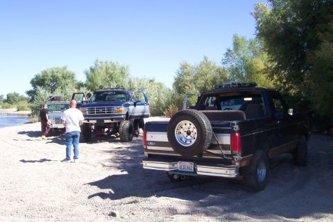 1995 Ford Bronco - Multiple lifted trucks are parked on a sandy area near a body of water.