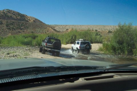 1995 Ford Bronco - Two SUVs navigate a dry riverbed under a clear blue sky, kicking up water as they go.
