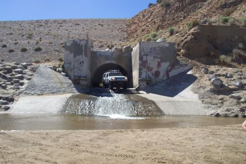 1995 Ford Bronco - A white SUV drives through a water channel exiting a concrete tunnel in a dry, hilly area.