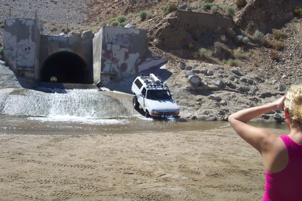 White SUV with roof rack driving through shallow water near a concrete tunnel