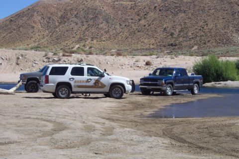 1995 Ford Bronco - A white sheriff SUV is parked next to two pickup trucks in a dry riverbed with a mountainous backdrop.
