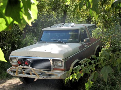 1978 Ford Bronco - A two-tone brown and white 1978 Ford Bronco is partially obscured by foliage, an aftermarket bumper and auxiliary lights.