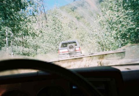 1991 Ford Bronco - An SUV navigates a dusty, wooded trail, viewed from the perspective of a driver behind it.