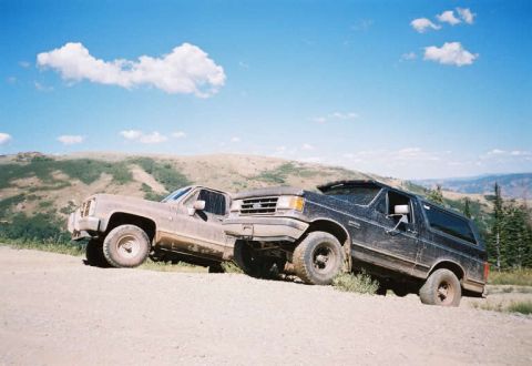 1991 Ford Bronco - Two SUVs, including a 1991 Ford Bronco, navigate a dusty mountain road under a bright blue sky.