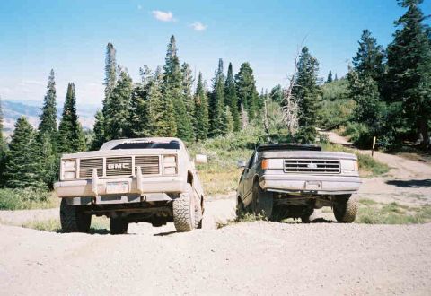 1991 Ford Bronco - Two trucks are parked on a dirt road in a mountainous, wooded area under a blue sky.