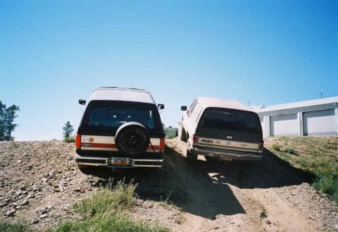 1991 Ford Bronco - Two SUVs, one a 1992 Ford Bronco, are parked on a dirt path with a building in the background under a clear blue sky.