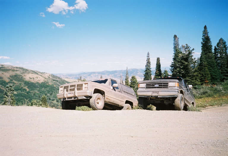 Two SUVs parked on a dirt road, one is a light brown older model, the other is a dark gray pickup truck