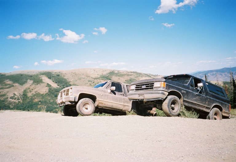 1991 Ford Bronco and an older pickup truck parked on a dirt road with hills in the background