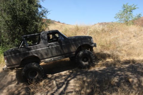 1992 Ford Bronco - A dark gray Ford Bronco with large off-road tires navigates a dusty, dry trail under a clear blue sky.
