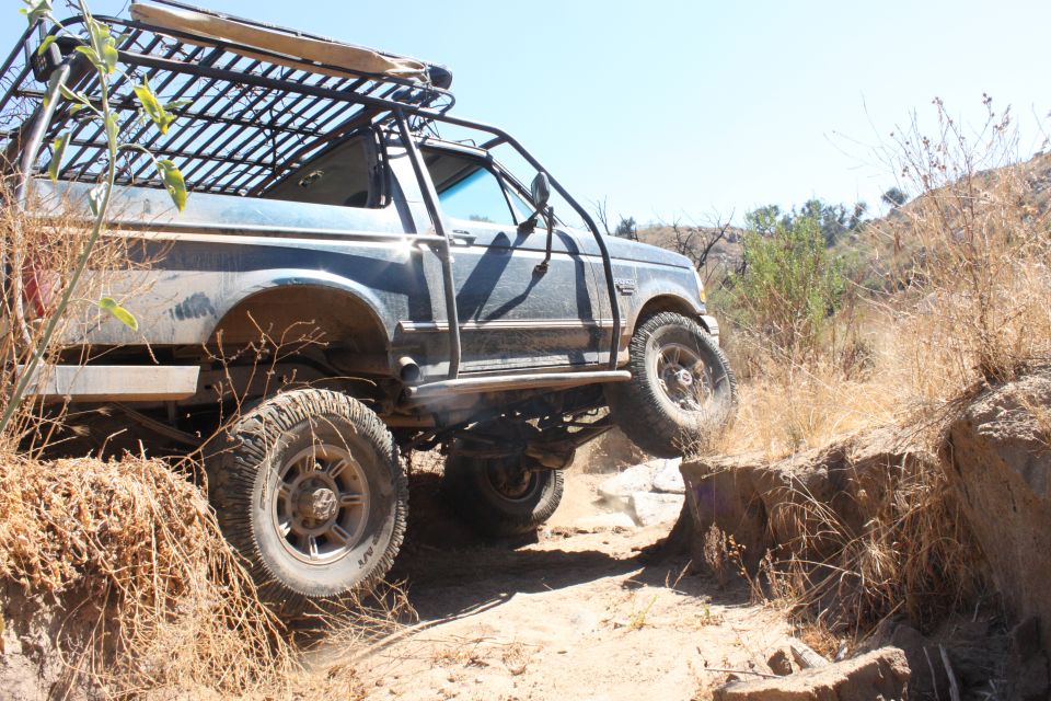 Ford Bronco with a roof rack and roll cage driving on a dirt trail, tires on rocks