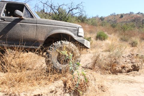 1992 Ford Bronco - This SUV is covered in mud while navigating a dry, brushy terrain with hills in the distance.