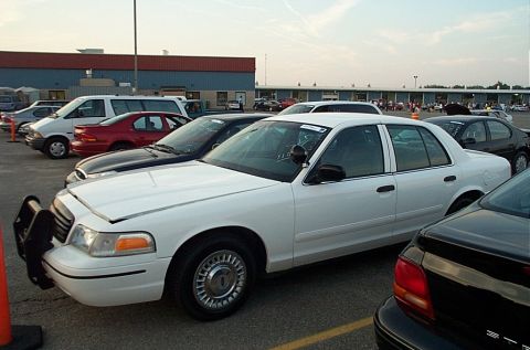 2001 Ford Crown Victoria - Side profile of a white Ford Crown Victoria police sedan in an auto auction lot, showing a black push bumper on the nose, a lifted hood-edg…