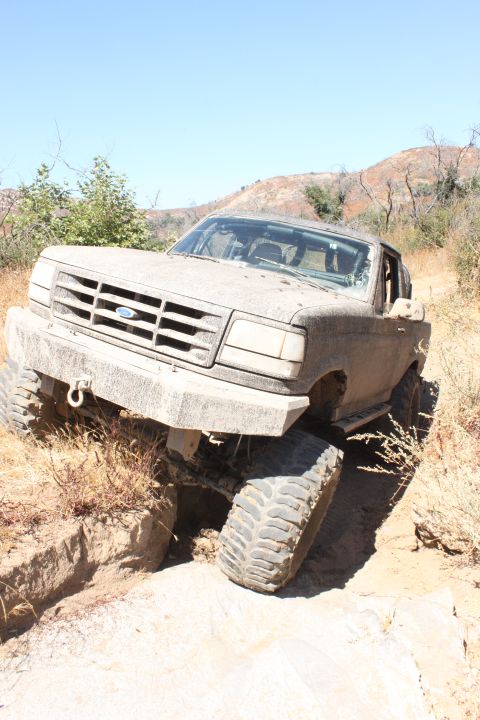 1992 Ford Bronco - A mud-covered 1992 Ford Bronco navigates a challenging dirt trail, with one front wheel suspended in the air.