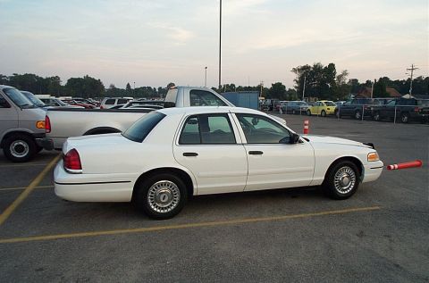 2001 Ford Crown Victoria - Rear three-quarter side view of a white Ford Crown Victoria sedan parked on a paved lot, wearing silver full wheel covers, body-color mirro…