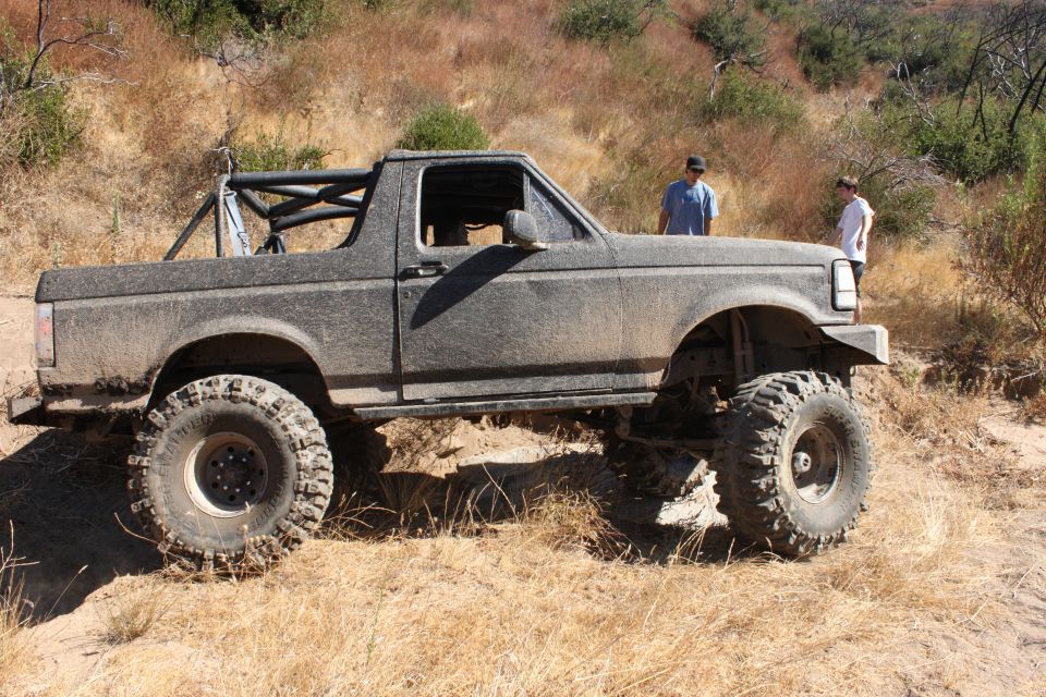 Mud-covered Ford Bronco with large off-road tires and roll cage parked on a dirt trail