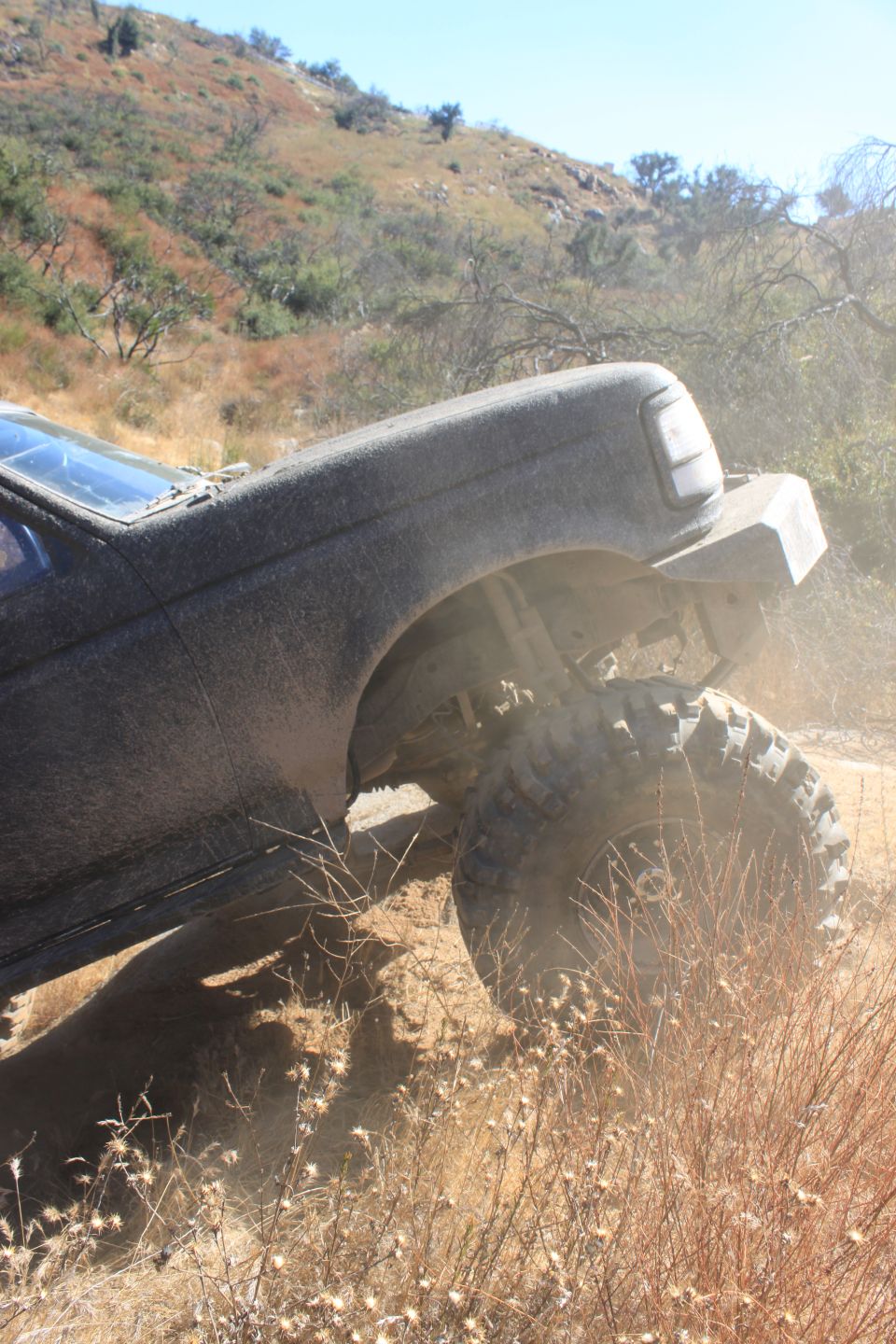 Mud-covered 1992 Ford Bronco on a dirt trail, viewed from the front 3/4 angle, showing its large tires and off-road modifications