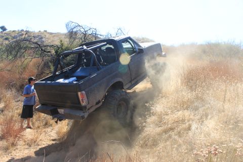 1992 Ford Bronco - A modified SUV kicks up dust as it ascends a steep, dry, grassy incline on a sunny day.