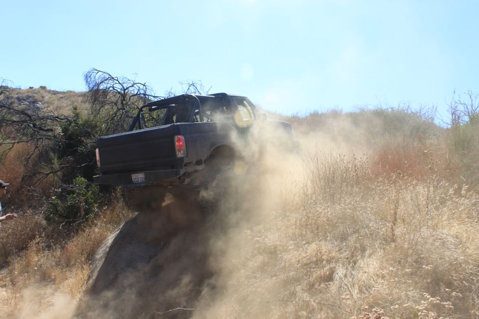 Black off-road vehicle kicking up dust while climbing a steep, rocky hill. A person stands to the left