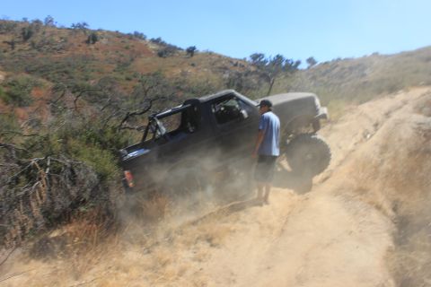 1992 Ford Bronco - A black off-road truck navigates a dusty, hilly terrain with a person standing nearby.