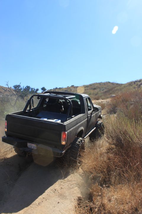 1992 Ford Bronco - A black 1992 Ford Bronco with a roll cage navigates a dusty dirt trail in a dry, hilly landscape.