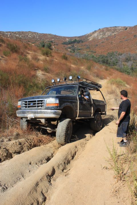 1992 Ford Bronco - A man stands by a lifted Ford F-Series truck navigating a rutted dirt path in a dry, hilly landscape.