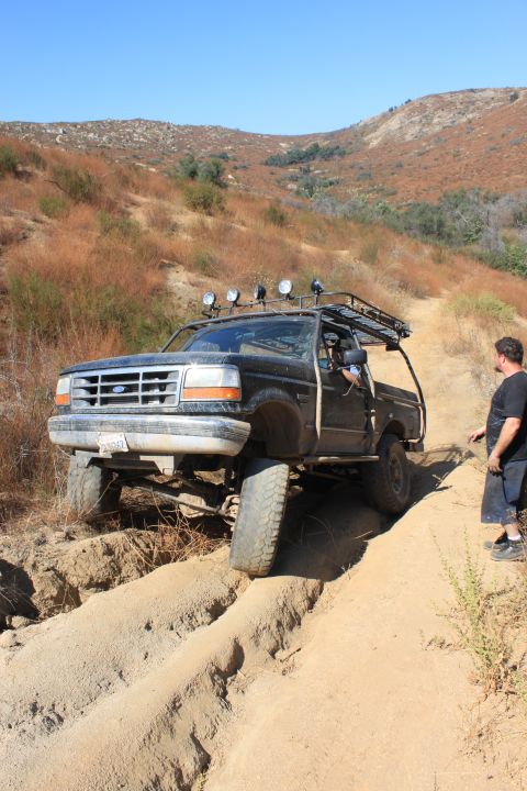 1992 Ford Bronco - A black 1992 Ford Bronco is shown off-roading on a dry, hilly terrain with a man standing nearby.