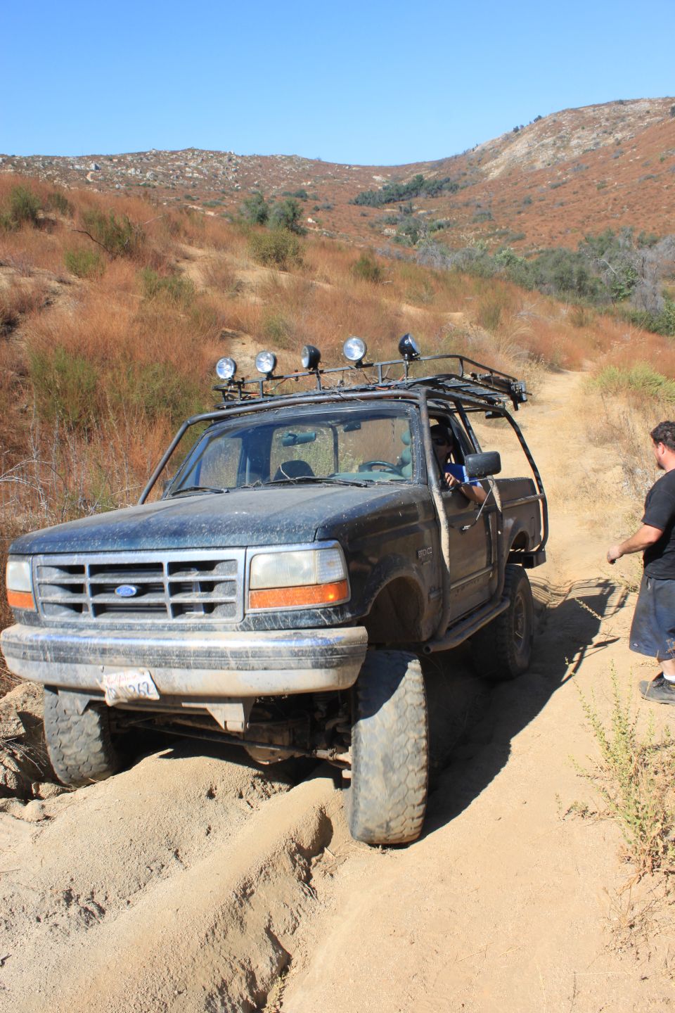 1992 Ford Bronco with roof rack and off-road lights navigating a sandy trail with a person standing nearby