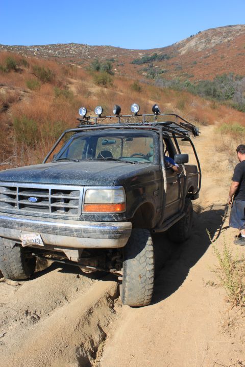 1992 Ford Bronco - This Ford Bronco is navigating a challenging off-road trail, with one wheel stuck in a deep rut.