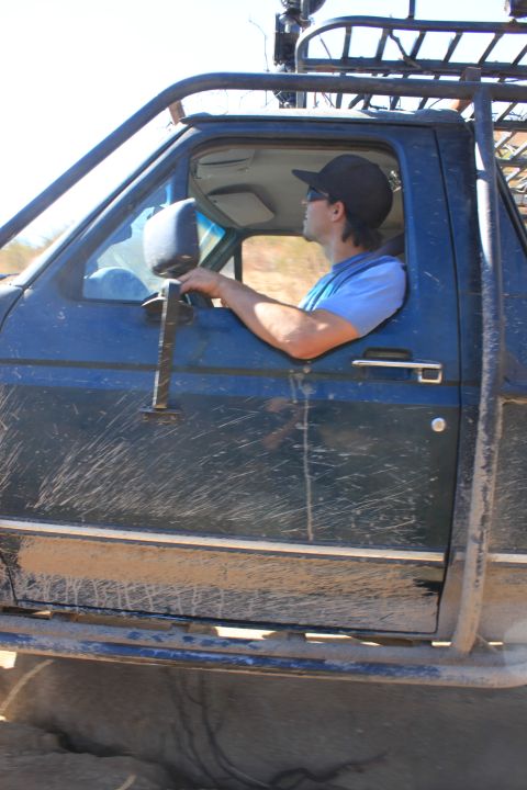 1992 Ford Bronco - Driver in a blue shirt and black cap steers a mud-splattered blue off-road truck on a trail.