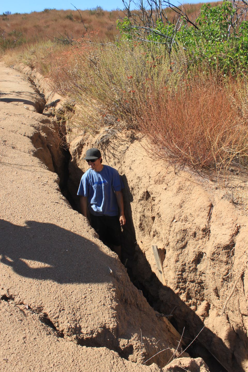 Man in blue shirt and black cap walks through a deep dirt trench with dry brush on the sides