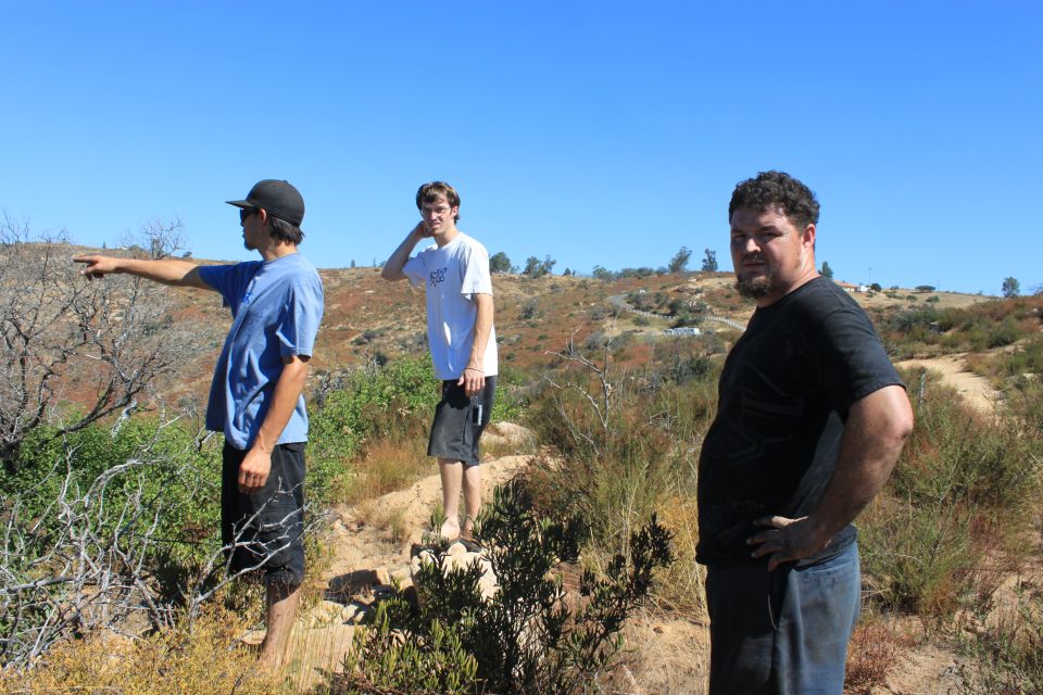 Three men stand in a dry, hilly landscape under a clear blue sky. One man points to the left