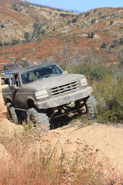 1992 Ford Bronco - Two off-road trucks navigate a dusty, hilly trail with dry vegetation.