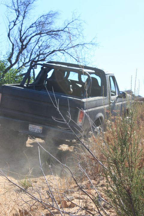 1992 Ford Bronco - A dark SUV with a roll cage navigates a dusty trail, seen from the rear.