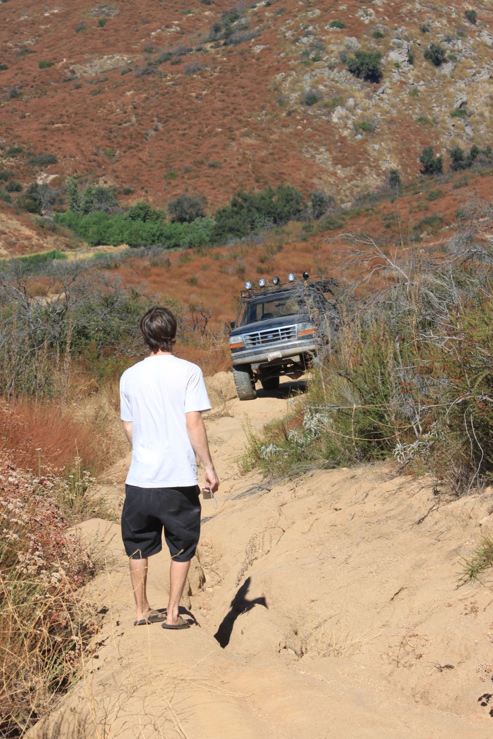 1992 Ford Bronco driving up a sandy, off-road trail with a person walking behind it