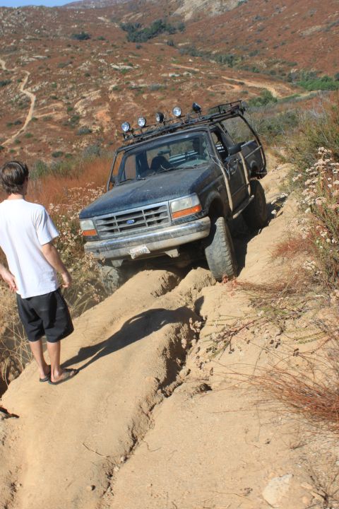 1992 Ford Bronco - A dark 1992 Ford Bronco navigates a challenging off-road trail, with a person observing from the side.