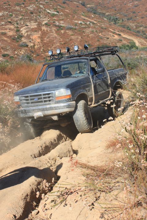 1992 Ford Bronco - A dark-colored Ford Bronco tackles a challenging off-road trail, kicking up dust as it goes.